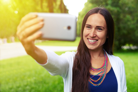 Close-up of beautiful woman posing at the camera on her smart phone making a selfie in the parkの写真素材