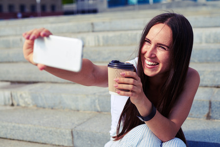 Attractive woman with long dark hair is making a selfie with a cup of coffee in her hands against stairs backgroundの写真素材