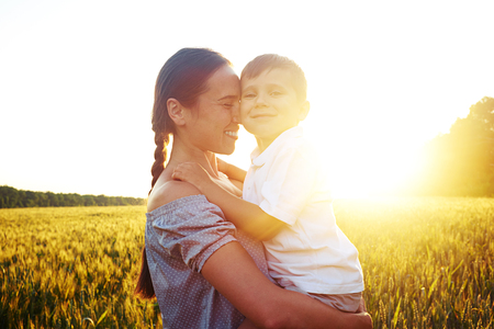 Young beautiful woman is holding small boy in arms against magnificent sunset in the fieldの写真素材