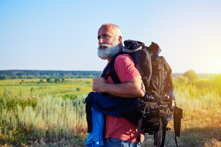 Fit handsome aged man with white beard in casual clothes is standing in front of sunlit field carring a rucksackの写真素材