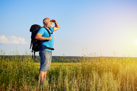 Physically fit handsome aged man with rucksack is standing in the grass and looking at sunlightの写真素材
