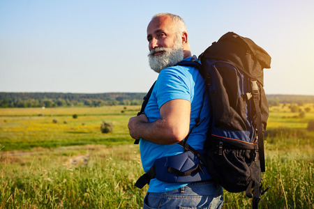Close-up of physically fit aged man with rucksack who turned his head to smile at the camera while walking in the field on sunny summer dayの写真素材
