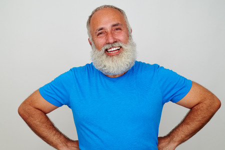 Handsome aged man in blue T-shirt is standing with hands on hips and smiling against white backgroundの写真素材