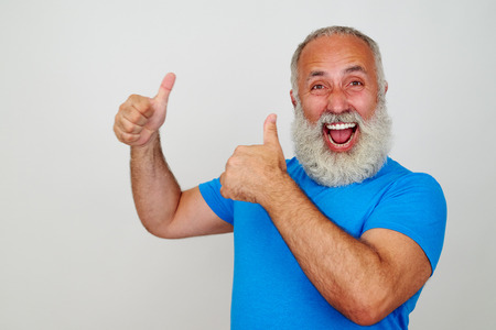 Smiling bearded aged man in bright blue T-shirt is showing two thumbs up gesture isolated against white backgroundの写真素材