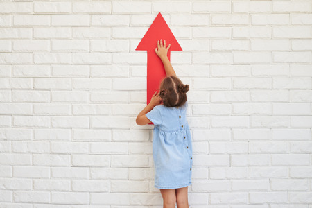 Pretty little kid in a light-blue dress holding a big arrow in hands over a brick white wallの写真素材