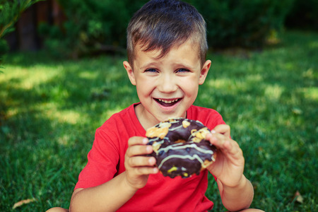 A handsome boy with cornflower blue eyes holding a doughnut and laughing, sitting on the grass and having good time in the parkの写真素材