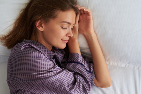 Close-up of serene beautiful woman sleeping on her left side on white pillow with hands under her...