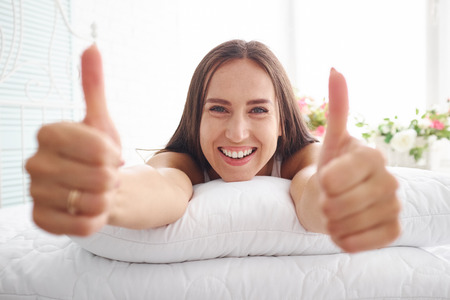 Portrait of beautiful woman lying on bed with broad smile with her hands resting on white cushion and showing two thumbs upの写真素材