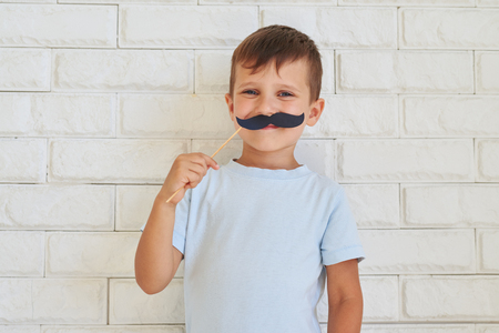 Young gentleman holding paper moustache with rather mischievous facial expression standing against white wallの写真素材