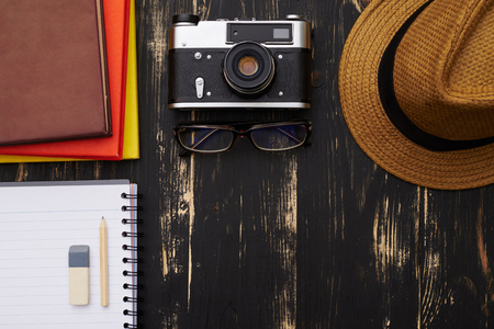 Top view of notepads, stationery objects, hat, vintage camera and eyeglasses on dark wooden desk backgroundの写真素材