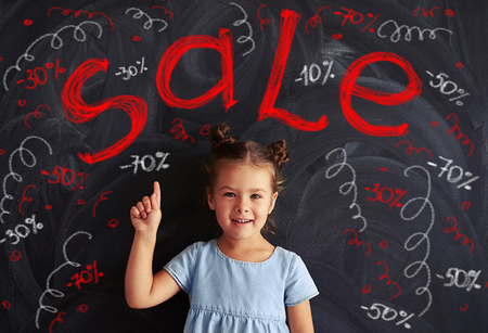 Small cute girl in a light-blue dress pointing with her finger at the blackboard with chalk inscriptions of sale themeの写真素材