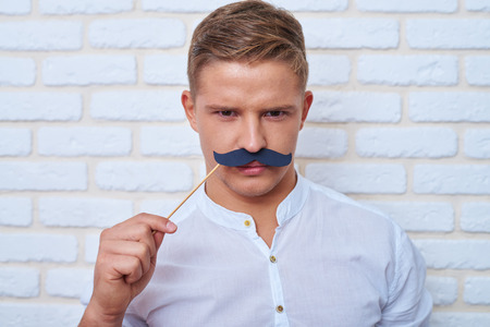 Close-up man looking at camera and smiling, standing against white brick wall in studio. Holding paper moustache and frowningの写真素材