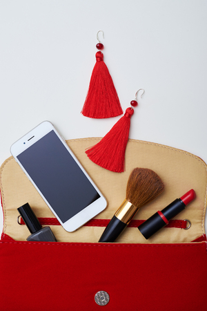 A close-up shot of bright reticule composed of brown makeup brush, big red earings, white mobile, lipstick and nail polish. Objects placed on light territoryの写真素材