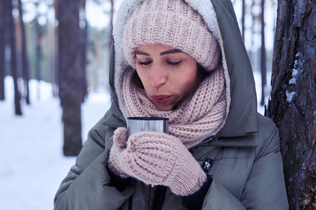 Close-up of woman in warm clothing blows on tea to cool the liquid down. Woman holding a hot drink. Model wearing padded jacket with knitted scarf and mittensの写真素材