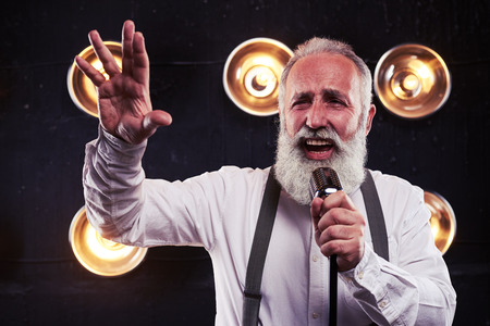 Close-up of emotional man in white shirt and suspenders holding a retro silver studio microphone over the background of blurred spotlightsの写真素材