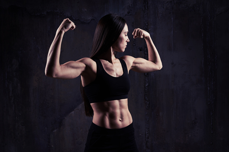 Close-up shot of woman flexing muscles showing great relief of arms and torso while exercising against rough dark wall with rusty gearsの写真素材