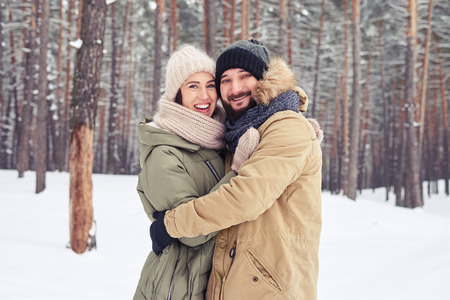 Close-up of loving couple standing in the woods. Smiling couple hugging each other and looking at the camera while standing in the wintry forestの写真素材