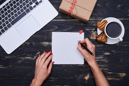 High angle of female hand writing a wish on a greeting card. Cup of coffee and biscuits, silver laptop isolated over grunge flat lay backgroundの写真素材