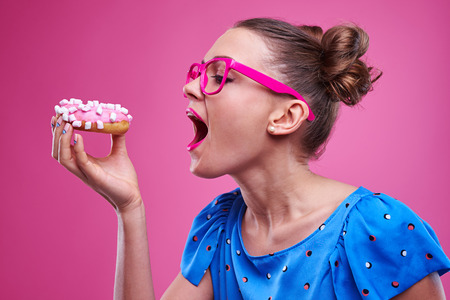 Side view of joyous young woman eating sprinkled doughnut isolated over pink background.の写真素材