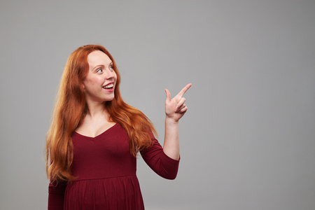 Mid shot of curious pregnant woman with red hair pointing to a copy space isolated over background in the studio. Promotion of somethingの写真素材