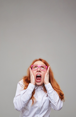 Close-up portrait of red hear teenager wearing pink glasses and white blouse looking frightened upwards. Place for a copy spaceの写真素材