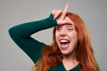 Close-up shot of young woman with loser sign on forehead isolated over gray background. Happy female having fun in the studioの写真素材