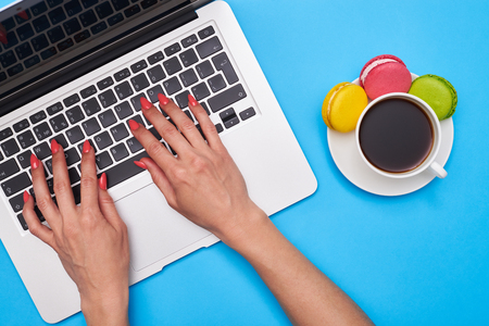 Top view of female hands pressing keys of black keyboard of the silver laptop. Delicious macaroons with a cup of coffee. Workplaceの写真素材