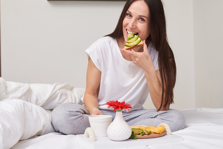 Mid shot of smiling woman in pajamas eating sandwich and enjoying romantic breakfast sitting in the bed with copy spaceの写真素材