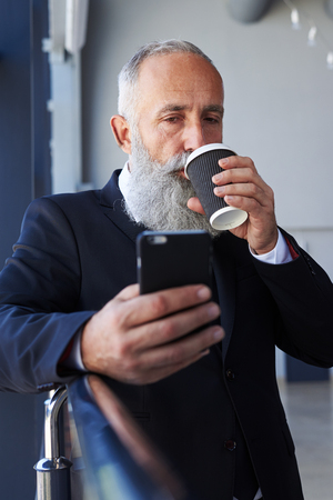 Vertical of bearded male age of 50-60 drinking coffee and surfing in phoneの写真素材