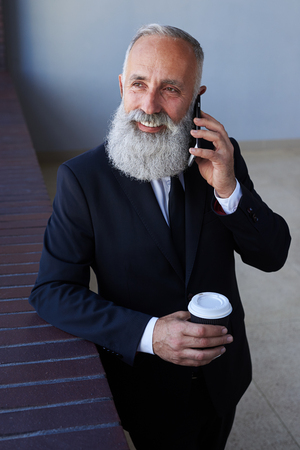 Vertical of cheerful gentleman talking on phone while holding cup of coffeeの写真素材