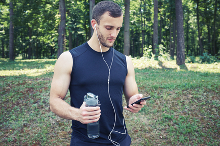 Mid shot of athlete watching something at mobile, wearing headphones in the street, holding a bottleの写真素材