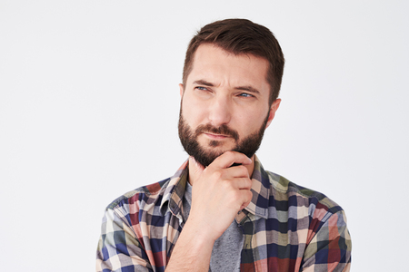 Close-up shot of young thoughtful man with beard holding hand on chin looking side thinking about something important. Isolated over white backgroundの写真素材