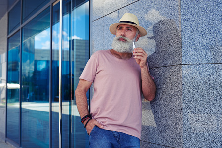 Mid shot of  bearded man with electro cigarette dressed in casual clothing, standing outdoors, narrowing eyes, holding hand in pocketの写真素材