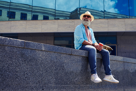 Smiling bearded man surrounded by skyscrapers, sitting, having cheerful appearance holding cup, mid...