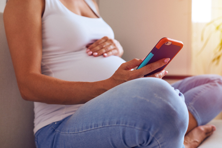 Cropped shot of hands of female touching pregnant belly and using phone at homeの写真素材