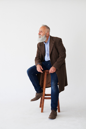 Full-length shot of candid mature bearded man in coat sitting on chair in studioの写真素材