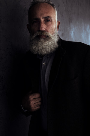 Closeup of bearded senior man in coat posing in dark studio background looking at cameraの写真素材