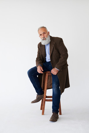 Full-length shot of serious mature bearded man in coat sitting on chair in studioの写真素材