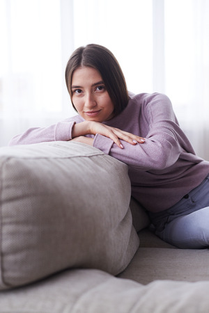 Vertical of happy young girl sitting on sofa in cosy clothsの写真素材