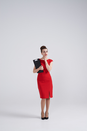 Full-length shot of woman in red dress holding hand on chin and looking thoughtfulの写真素材