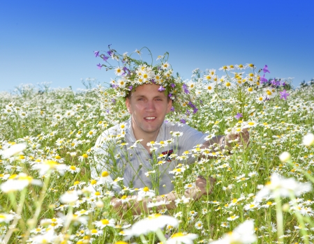 The smiling man in a wreath from wild flowers in the field of camomiles with a bouquetの写真素材