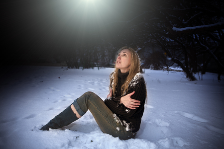 The young beautiful frozen woman sits in snow in the night parkの写真素材