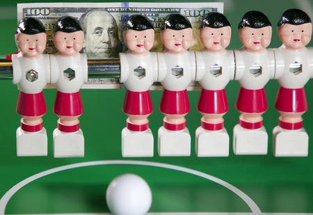 Sports and money concept -toy football players stand in the field and the banknote in hundred dollars on a backgroundの写真素材