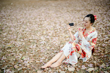 Cheerful Asian woman in floral kimono sitting on dry leaf ground, pointing at black card, traditional Japanese outfit and natural outdoor settingの写真素材