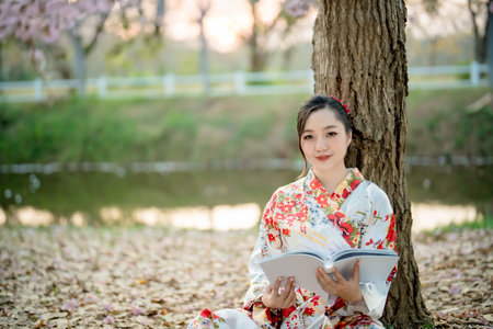 Asian woman in traditional floral kimono reading book under tree by the riverside in peaceful spring nature, enjoying calm moment of learning and relaxationの写真素材