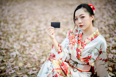 Beautiful Asian woman in traditional floral kimono holding black card while sitting on autumn leaf...