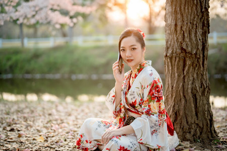 Elegant Asian woman in traditional floral kimono talking on smartphone under tree during sunset, blending Japanese heritage with modern digital lifestyleの写真素材