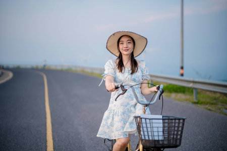Young woman in a summer dress and wide-brim hat riding a vintage bicycle on a scenic road with clear blue sky, enjoying peaceful outdoor lifestyle.の写真素材
