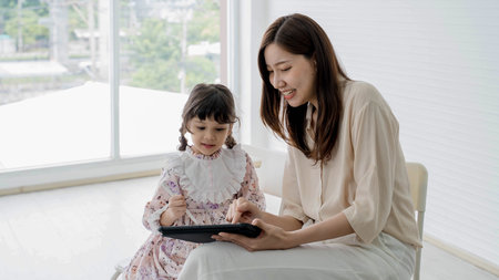 Mother and daughter using digital tablet together with stylus, smiling and learning happily at home in bright room, sharing fun screen time and creativity.の写真素材