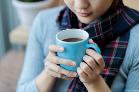 Close-up of woman gently holding a blue coffee mug while wearing a cozy plaid scarf, enjoying a moment of calm and warmth in a relaxed indoor environmentの写真素材